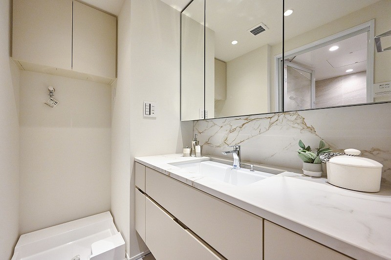 洗面室には室内洗濯機置き場と田中工藝製の洗面台があります。三面鏡裏や足元の収納棚、洗濯機置き場上の吊戸棚に収納スペースを作り、収納性を高めております。ゆとりのある広さの洗面台が出発前の身支度を捗らせてくれます。